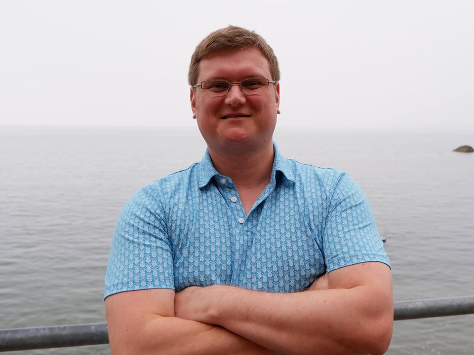 Graham at a seafront railing, arms folded, in front of a flat grey sea.
