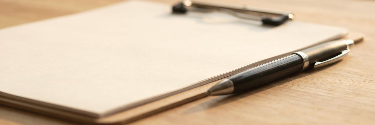 A clipboard holding blank printed sheets on a workbench, a ballpoint pen clipped to it.
