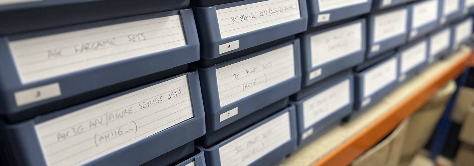 Rows of blue warehouse trays with handwritten labels, sorting paint and figure sets by range.